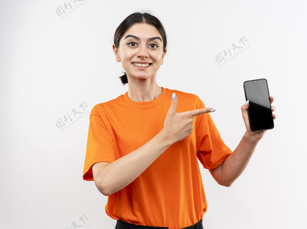 展示穿着橙色t恤的年轻女孩站在白色的墙上 用食指指着智能手机 开心地笑着女孩手指手机
