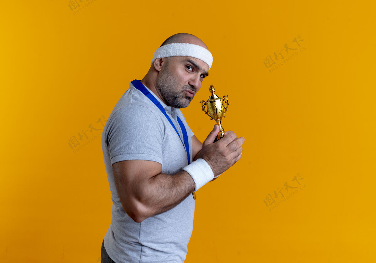 金牌戴着头巾 脖子上戴着金牌的成熟运动型男子拿着奖杯望着前方 严肃的脸站在橙色的墙上运动奖杯运动