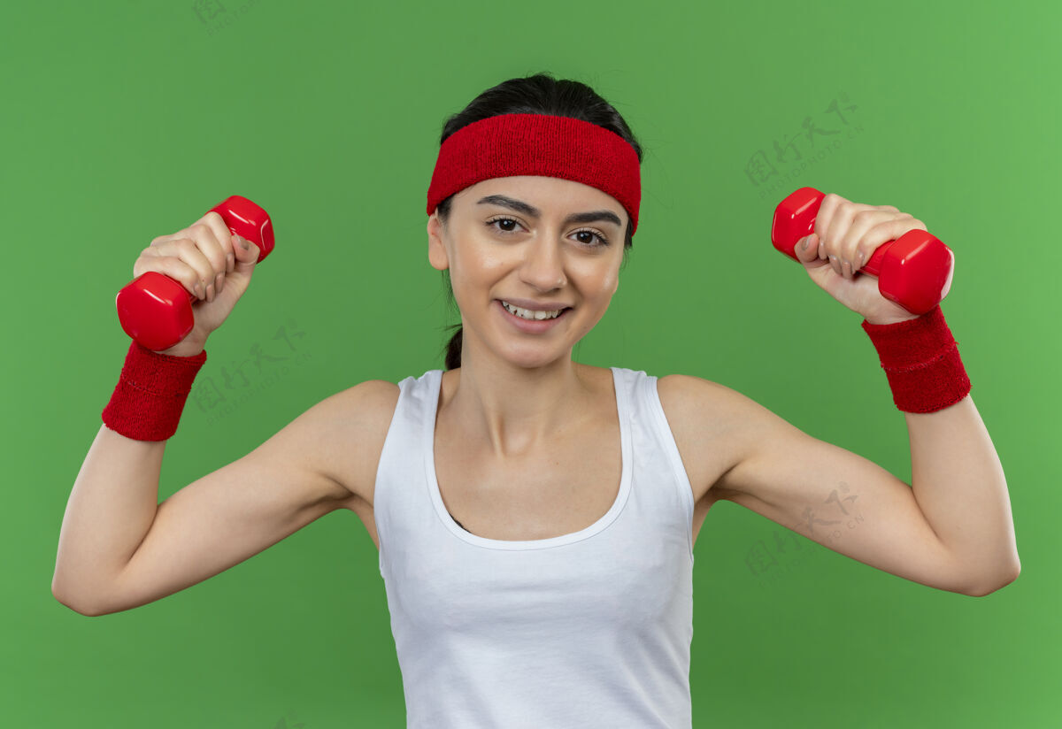 哑铃身穿运动服的年轻健身女士 头戴钢带 手持两个哑铃 举手练习 微笑自信地站在绿色的墙上自信看提高