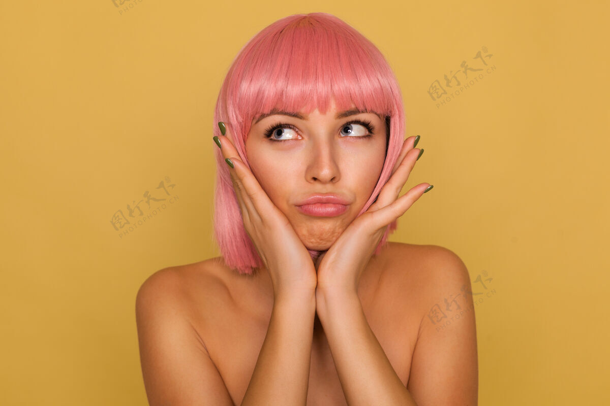爱情室内拍摄的年轻漂亮的蓝眼睛的女士 粉红色的鲍勃发型举着她的脸与提高手掌 向上看与困惑的脸 在芥末墙摆姿势短裤女人心情