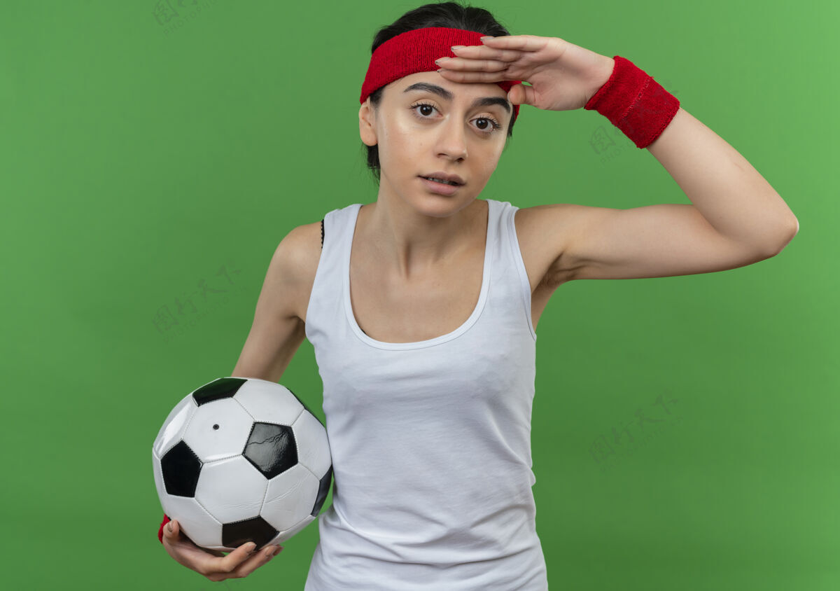 抱身穿运动服的年轻健身女 头戴足球带 手举着足球远远望着绿色的墙壁健康健身看