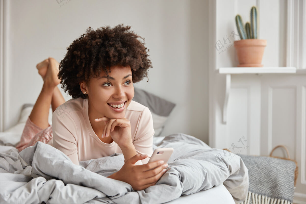 满意放松的黑皮肤女性 非洲发型 抬腿 穿着睡衣 手持现代手机 有梦幻般的表情 浏览互联网 发短信 享受平静的氛围女人室内非洲