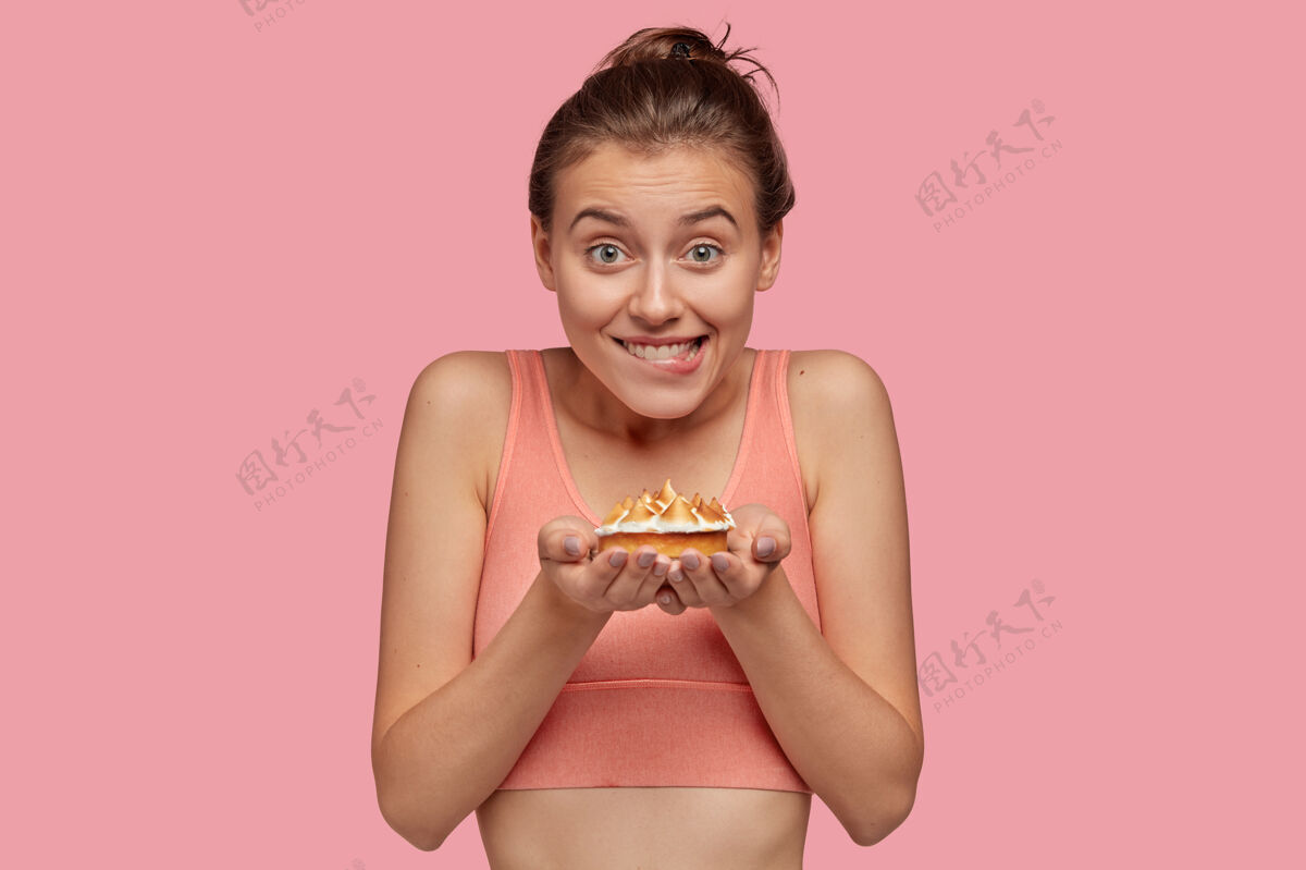 身体开朗健身女士的照片手捧美味甜饼 穿着休闲上衣 咬着嘴唇 不吃甜点想保持好身材 隔着粉色的墙 感觉诱惑肌肉菜品健身房