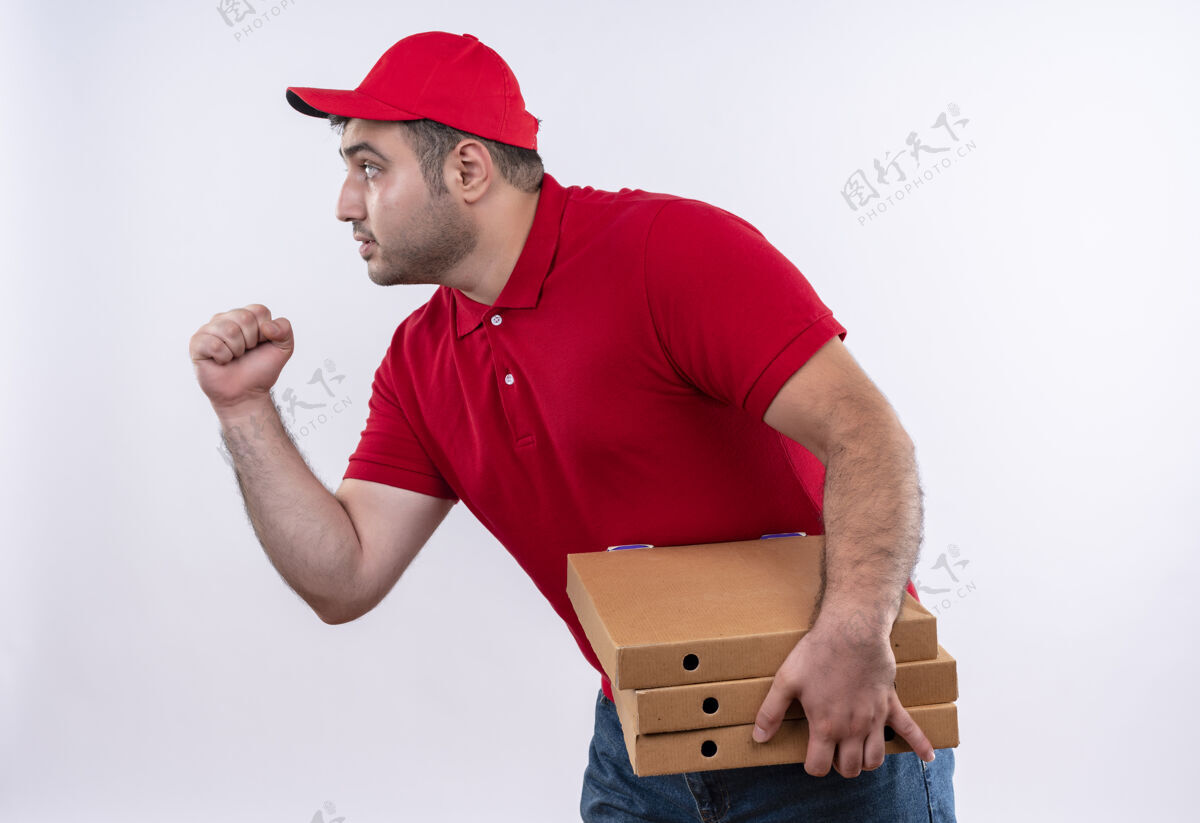 帽子年轻的送货员身穿红色制服 戴着帽子 手里拿着披萨盒 握紧拳头 面带严肃地站在白墙上盒子披萨年轻
