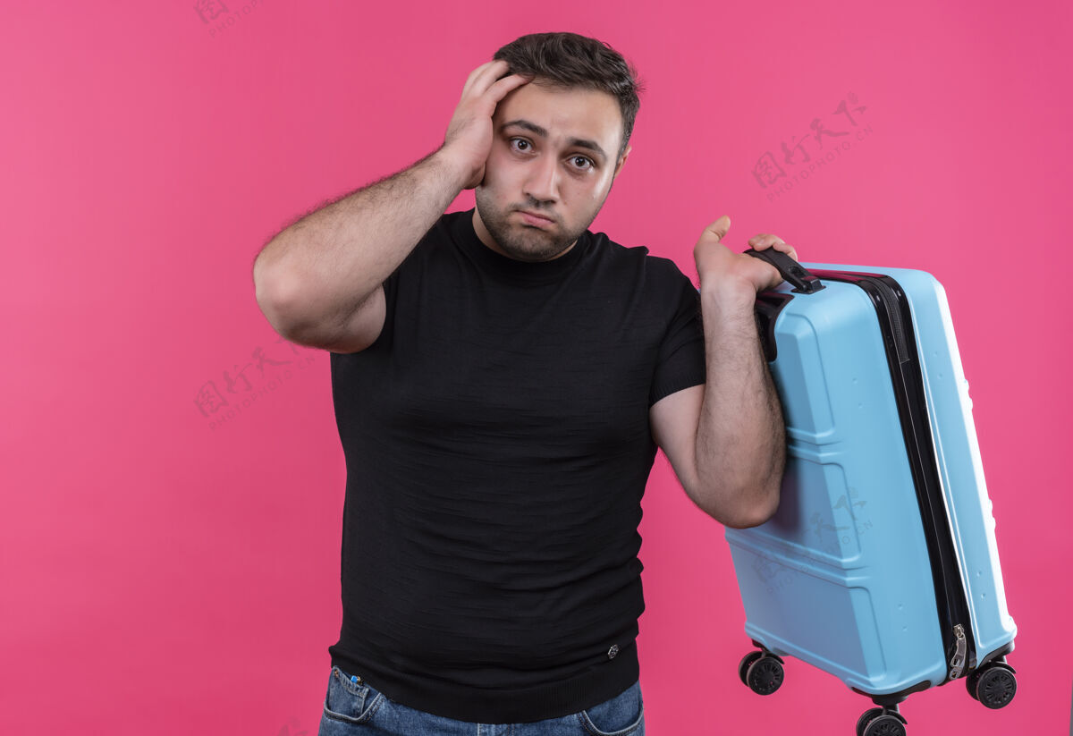 人穿着黑色t恤的年轻旅行家手里拿着手提箱 站在粉红色的墙上感到困惑和焦虑旅行者焦虑手提箱