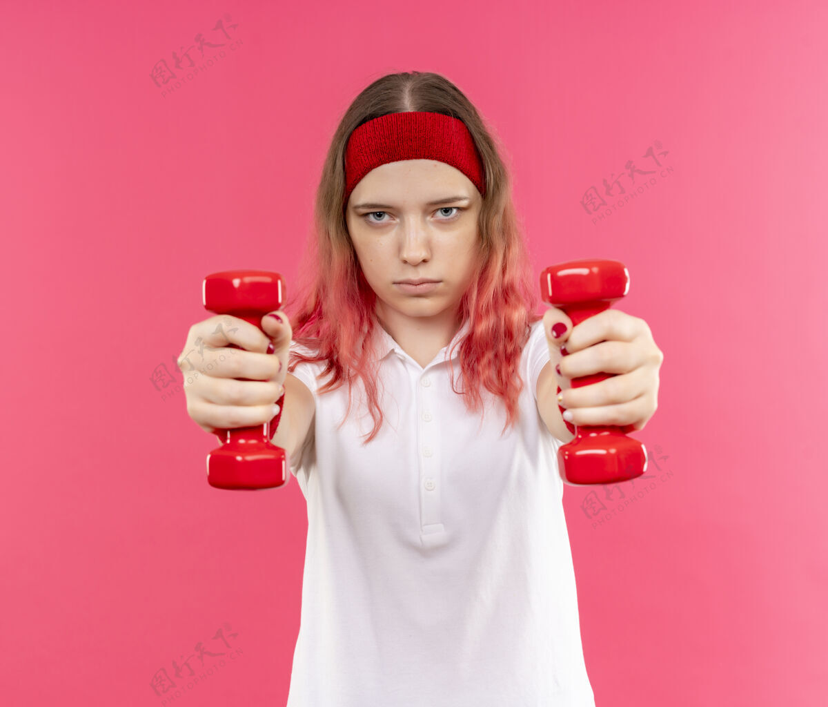 姿势戴着头巾的年轻运动女性站在粉色的墙上 用哑铃练习 表情严肃自信运动运动运动装
