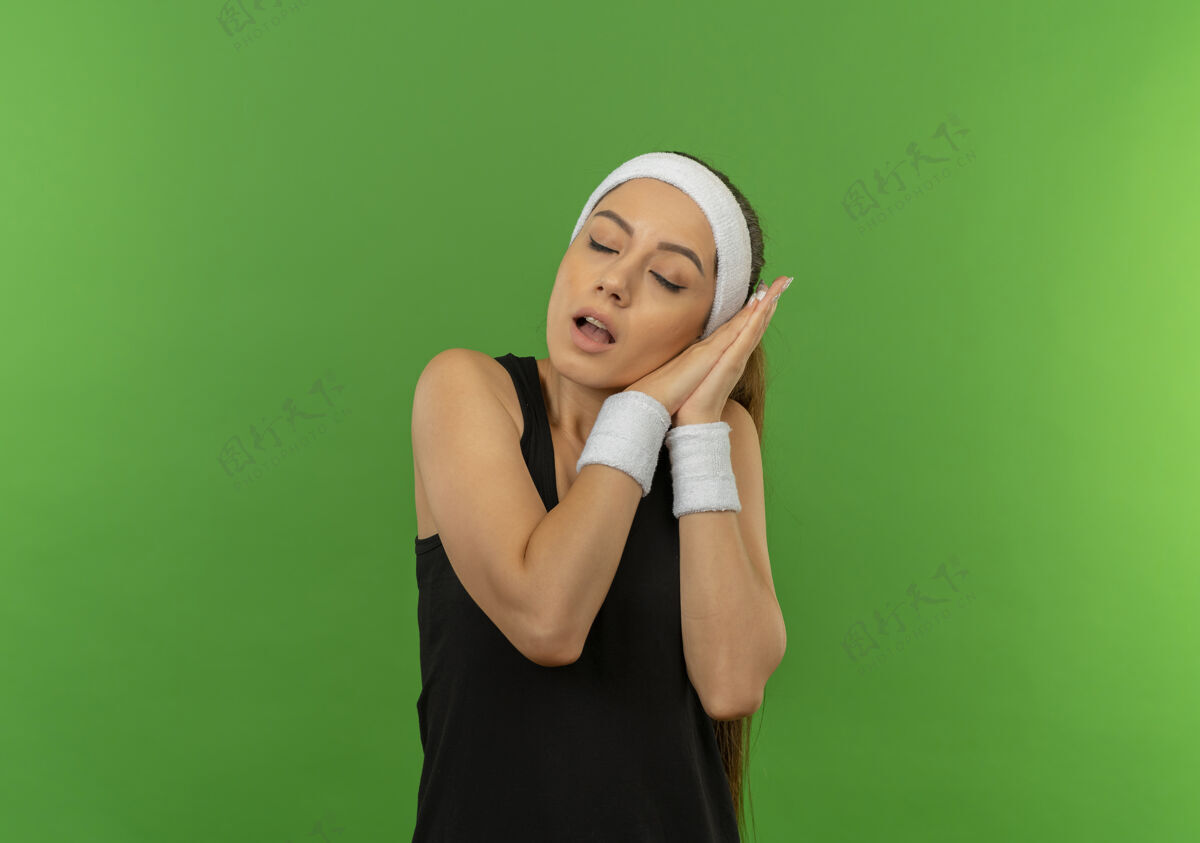 运动穿着运动服的年轻健身女士 头箍紧着手心 闭着眼睛 头靠着手心 想站在绿色的墙壁上睡觉抱着姿势头带