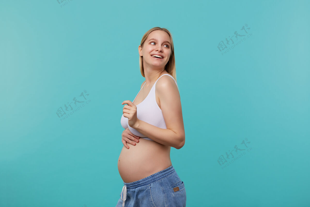 女士侧视图年轻迷人的白头未来的母亲看着幸福地在她的肩膀上 并举行了她的肚子 在蓝色背景孤立的手腹部怀孕金发