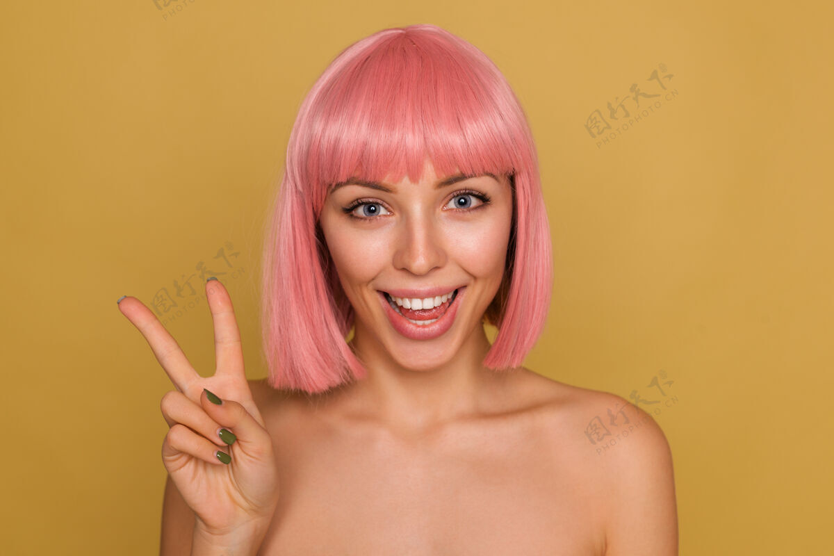欧洲室内拍摄迷人的年轻蓝眼睛粉红色头发的快乐女子举起手与胜利的姿态和微笑 而在芥末墙摆姿势提高发型手