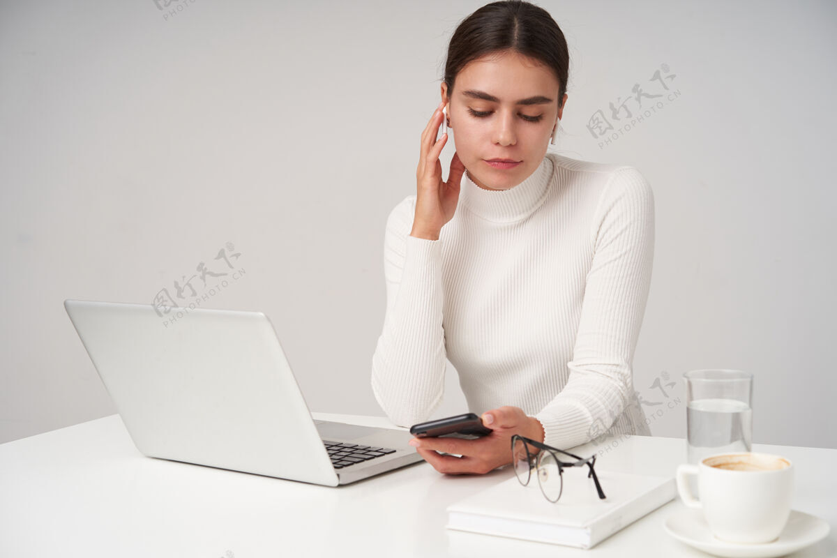 Poloneck室内照片 年轻漂亮的黑发女商人举起手对着耳机 手里拿着手机 坐在白墙上的桌子旁 手里拿着现代化的笔记本电脑心情马尾20多岁