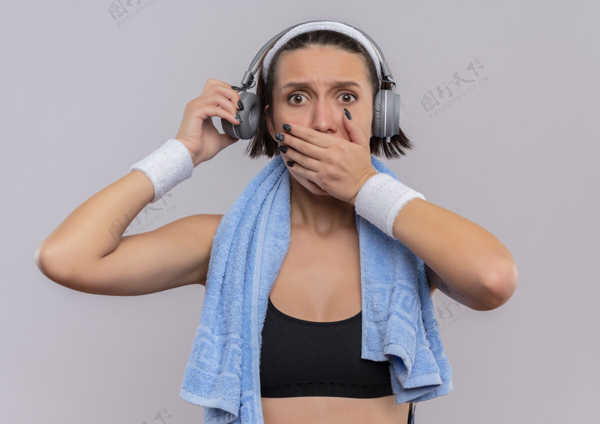 运动员一位身穿运动服 头戴耳机 脖子上套着毛巾的年轻健身女士用手捂住嘴巴 站在白墙上运动姿势女