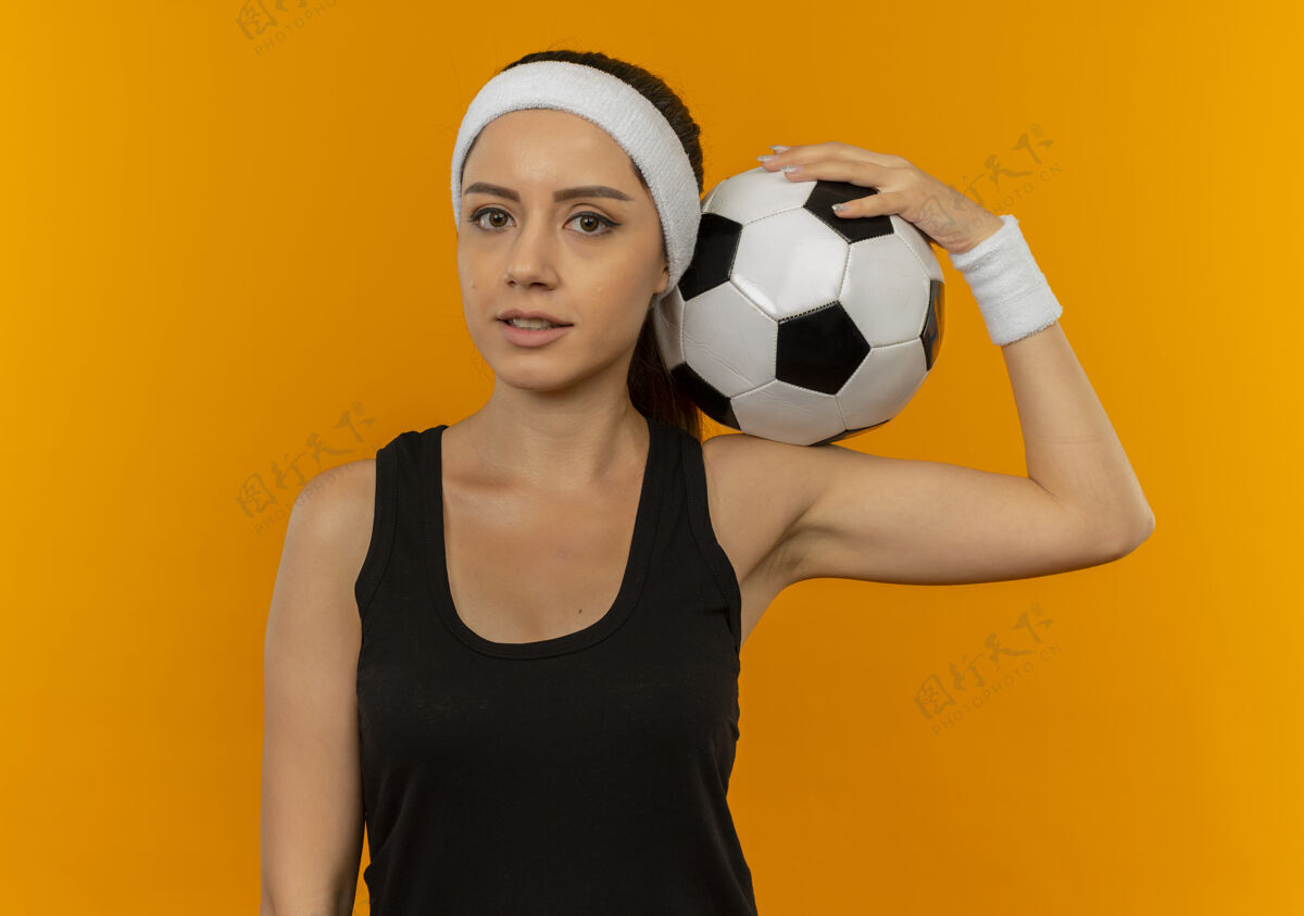 运动身穿运动服 头箍 手持足球的年轻健身女士站在橙色的墙上 看起来很自信公民运动装站着