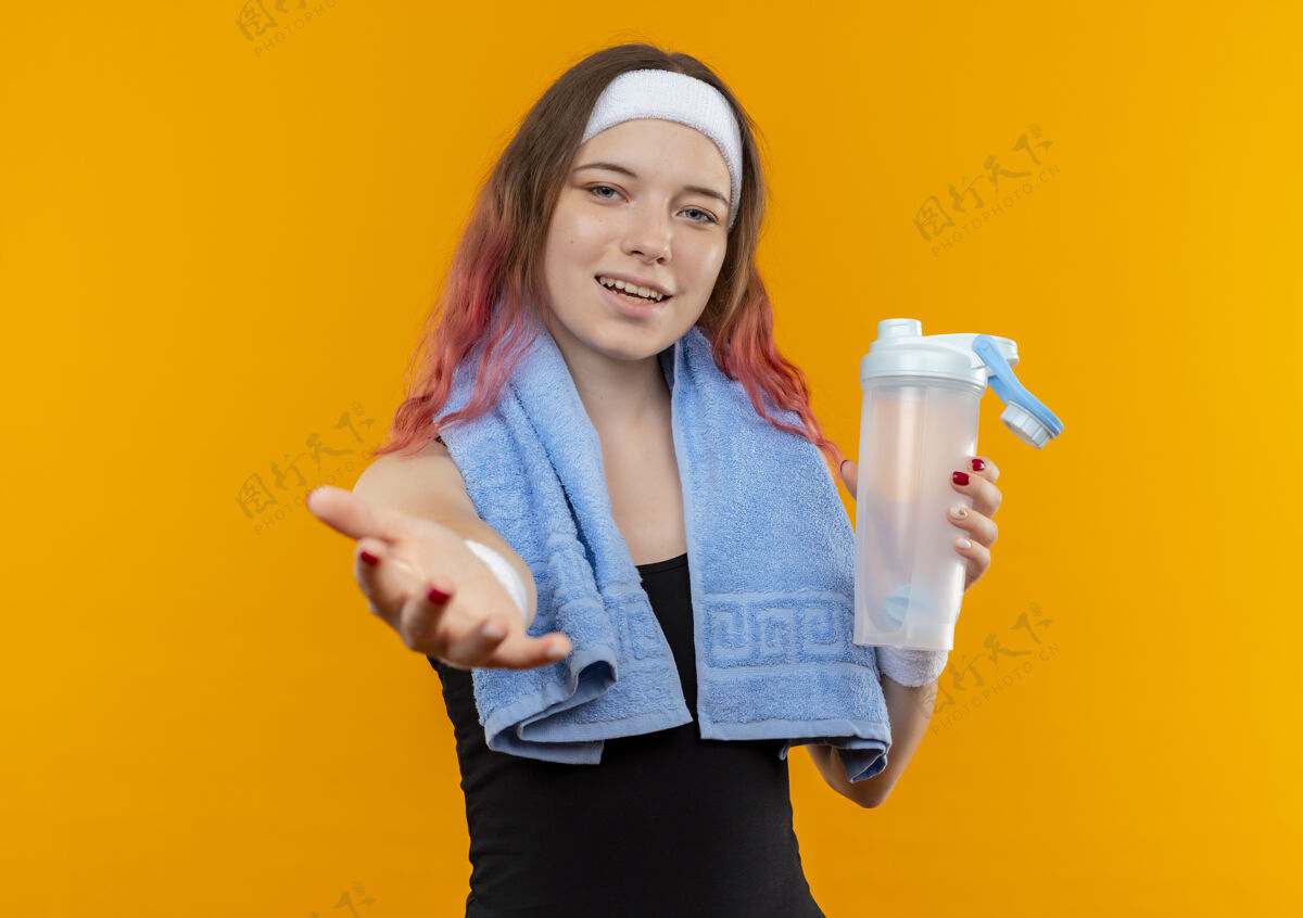 脖子身穿运动服的年轻健身女孩 脖子上套着毛巾 手里拿着装水的瓶子 站在橘色的墙上 手上露出愉快的微笑微笑健身运动装