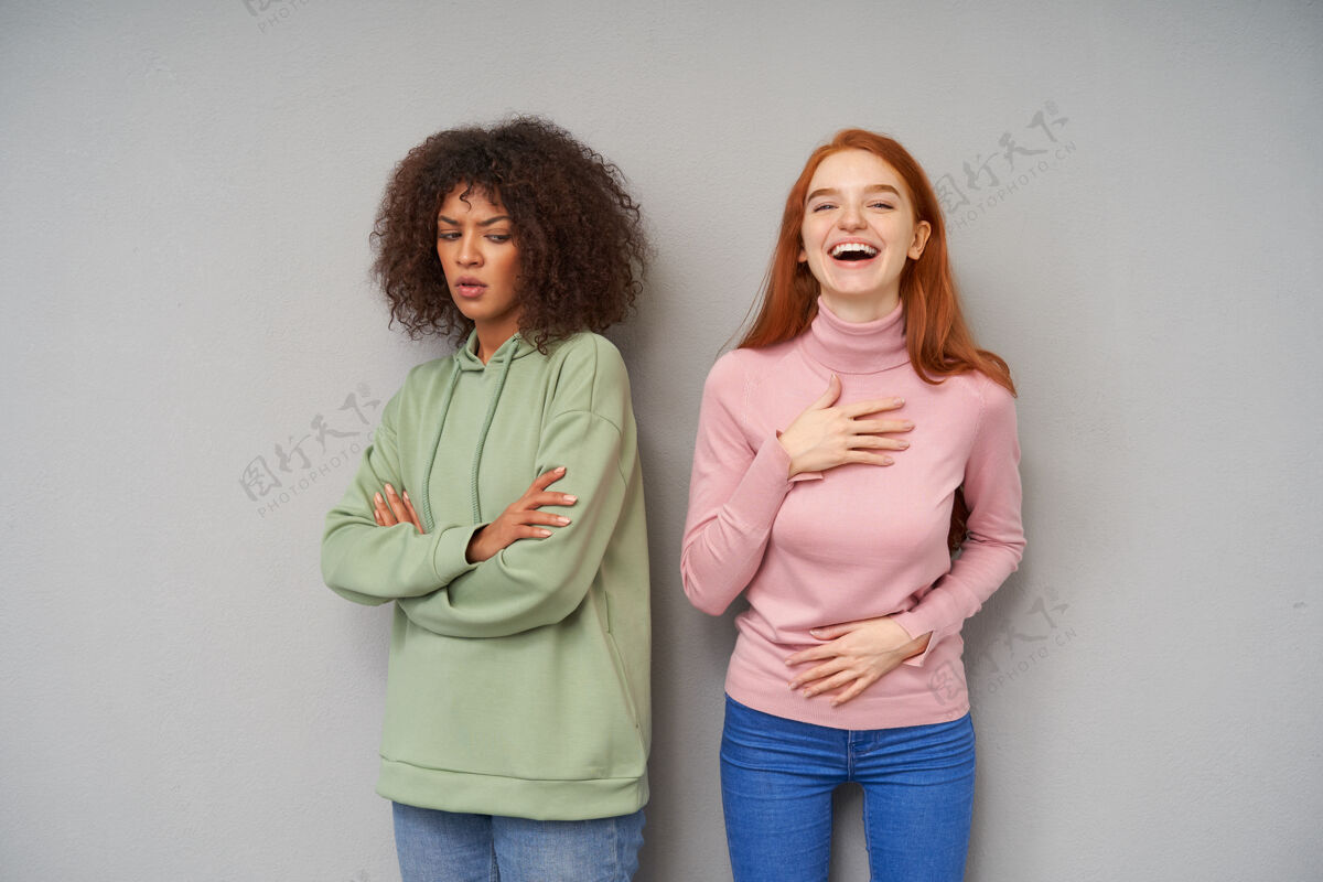 运动严肃的年轻棕发黑皮肤女人 双手合十 严肃地俯视着 一边在灰色的墙上摆姿势 一边是穿着休闲服的快乐的红发长发女人发型衣服爱