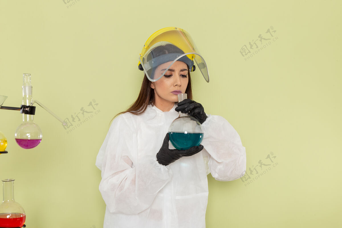 专业正面图穿着特殊防护服的女化学家拿着装有蓝色溶液的烧瓶 闻着绿色的表面化学家气味容器