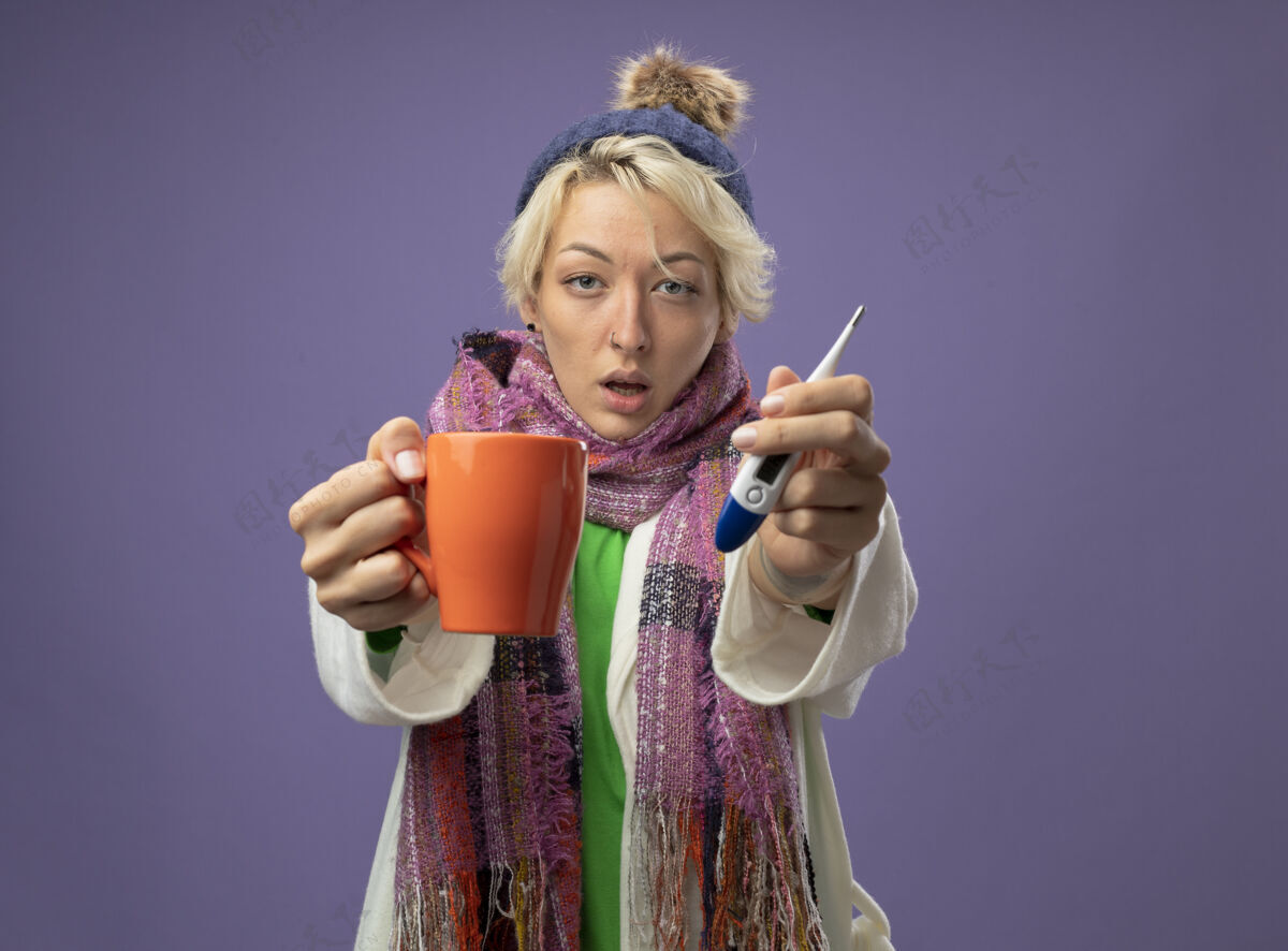 帽子有病的不健康的女人 短头发 戴着暖和的围巾和帽子 感觉不舒服 拿着一杯热茶和温度计 站在紫色背景上看着摄像机体温计不健康热