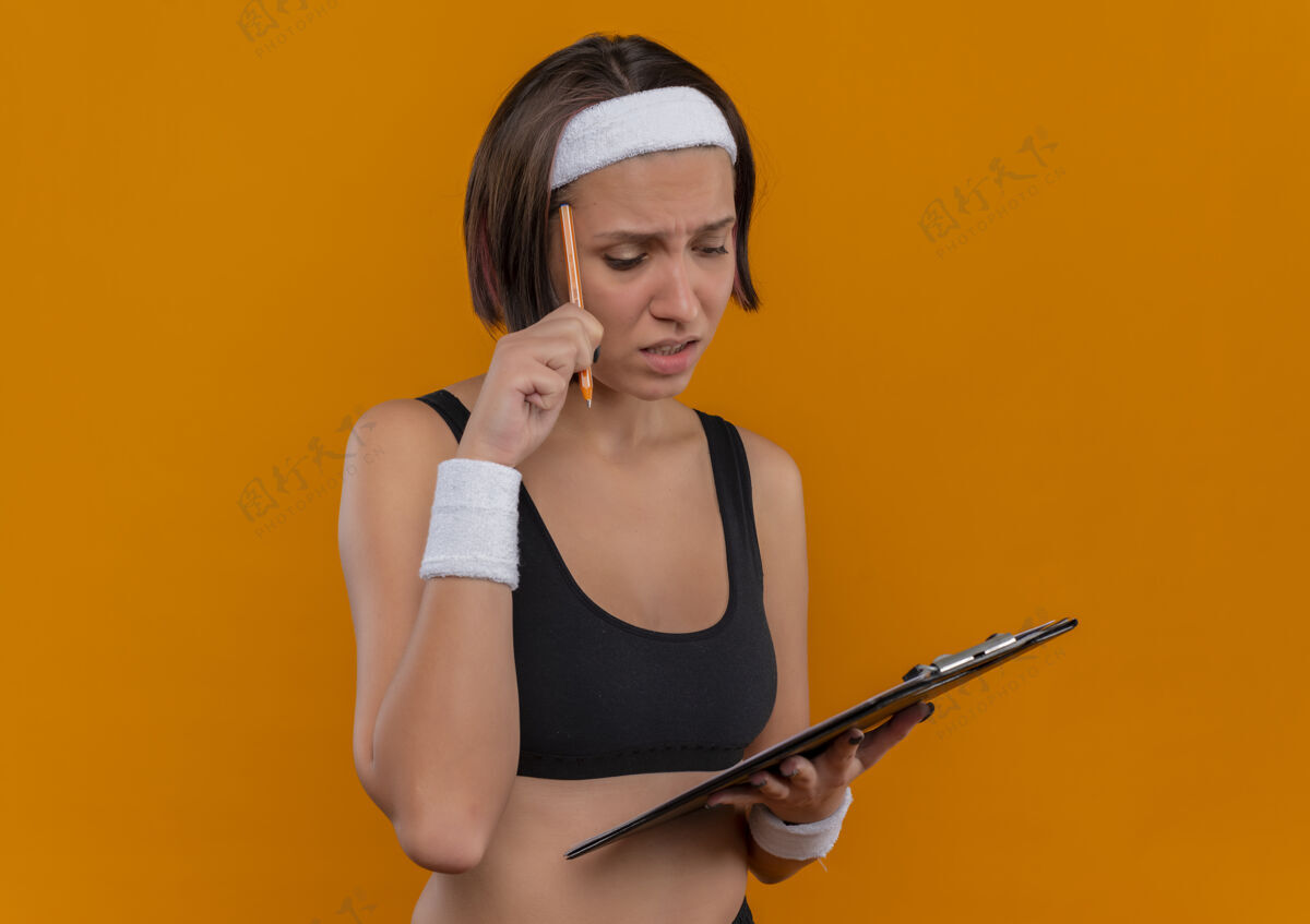 运动员穿着运动服的年轻健身女 戴着头巾 拿着空白页的剪贴板 站在橘色的墙上 用钢笔搔头 看起来很困惑公民运动装运动