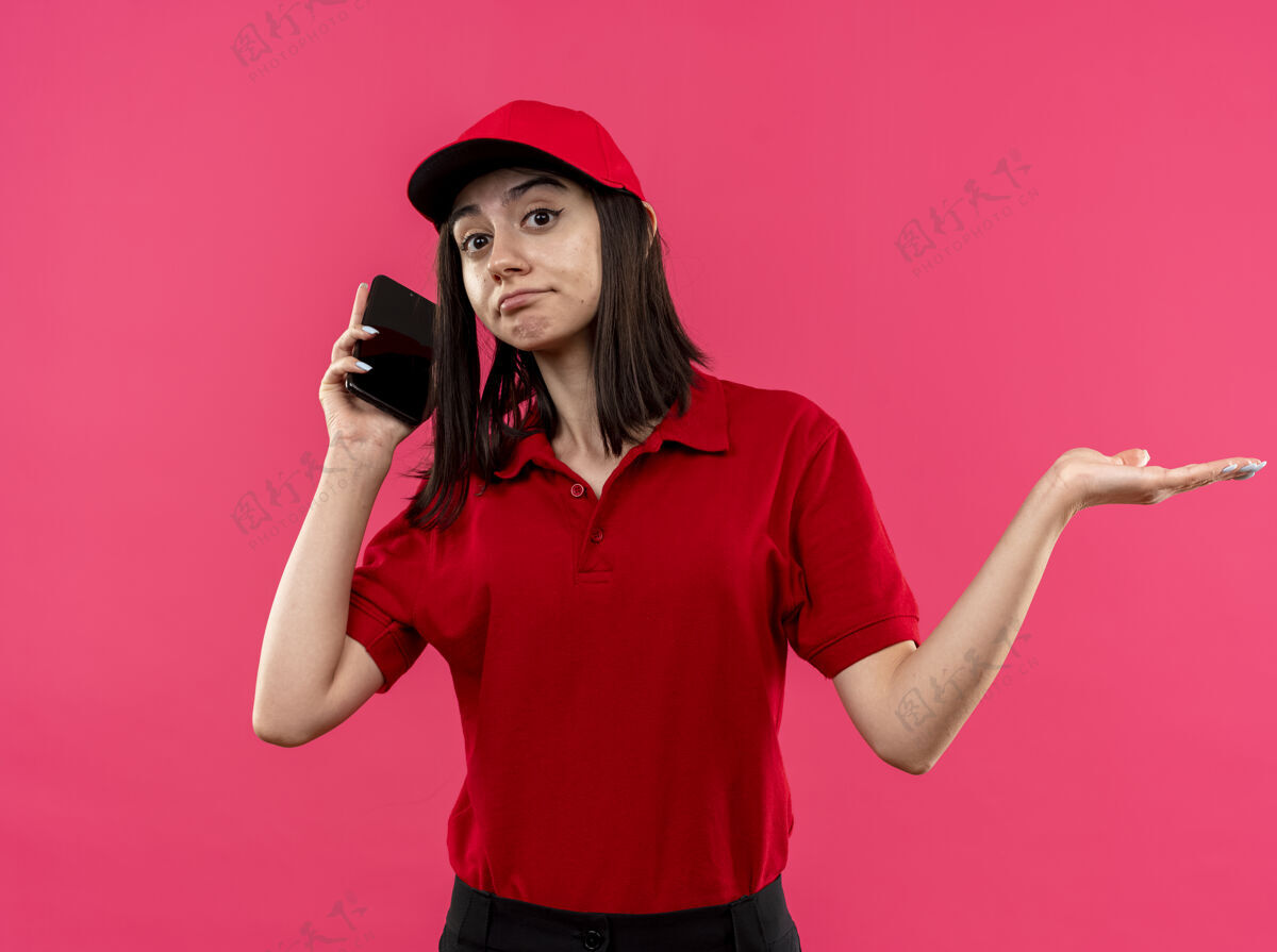 手机穿着红色马球衫和帽子的年轻送货女孩站在粉色背景下 一边说着手机 一边困惑地将手臂伸向一边马球困惑衬衫