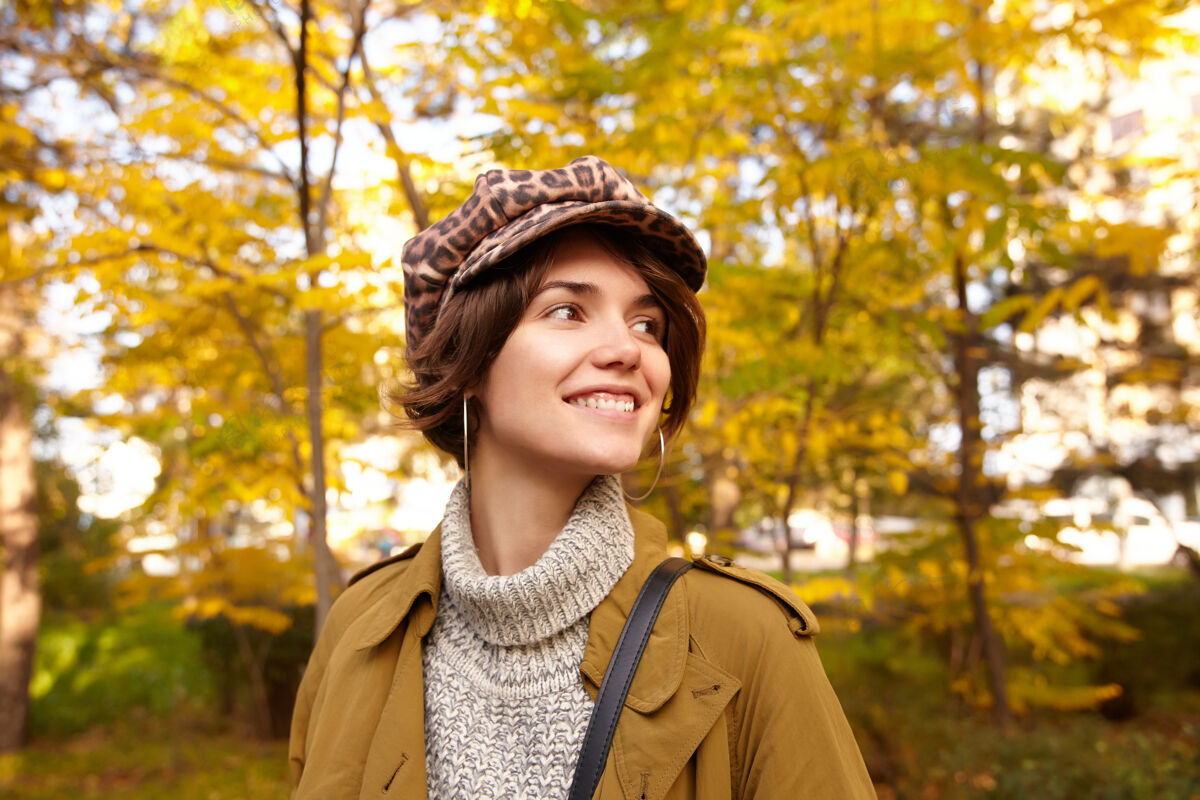 欢呼有吸引力的积极的棕色头发的年轻女子的特写与自然的化妆看一边 微笑着 而在时尚的穿着模糊公园摆姿势帽子城市海沟
