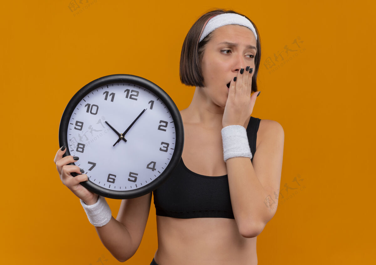人穿着运动服的年轻健身女士 头戴着挂钟 站在橘色的墙上打哈欠 看起来又累又无聊女性哈欠姿势