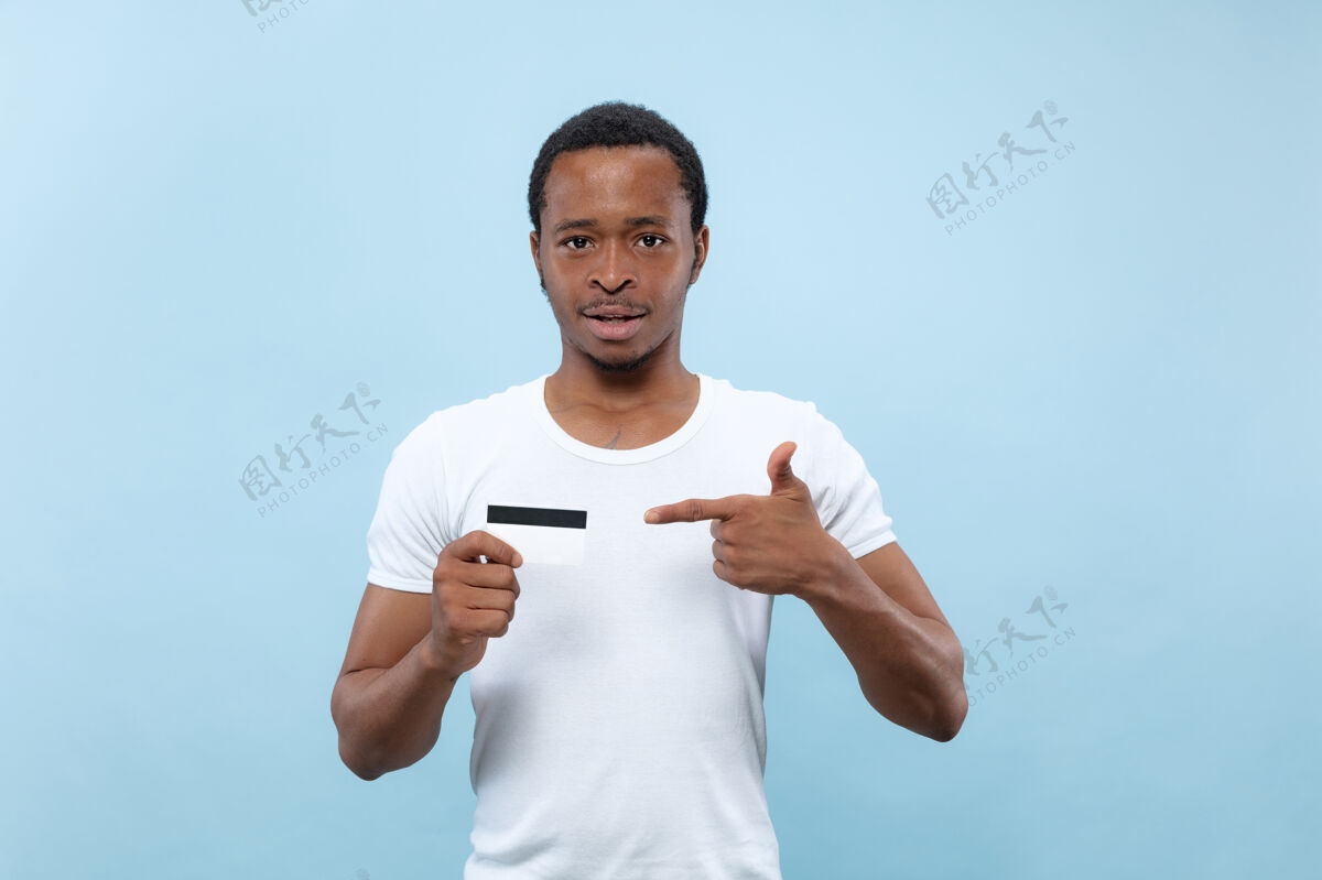 长相半身特写 蓝色背景 身穿白衬衫的非洲裔美国年轻男子人类情感 面部表情 广告 销售理念指向卡片付款 财务 账单非洲公司男性
