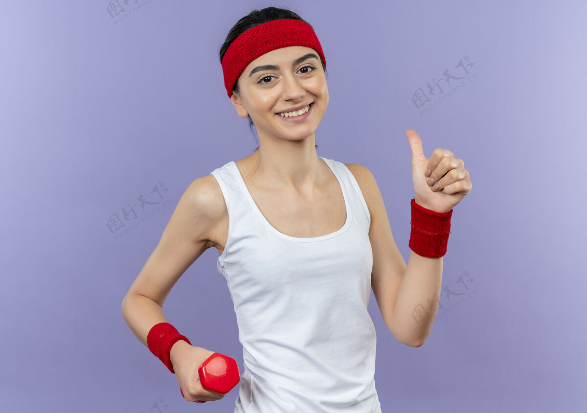 健身身穿运动服 头箍微笑的年轻健身女士站在紫色的墙上 愉快地竖起大拇指站立女人姿势