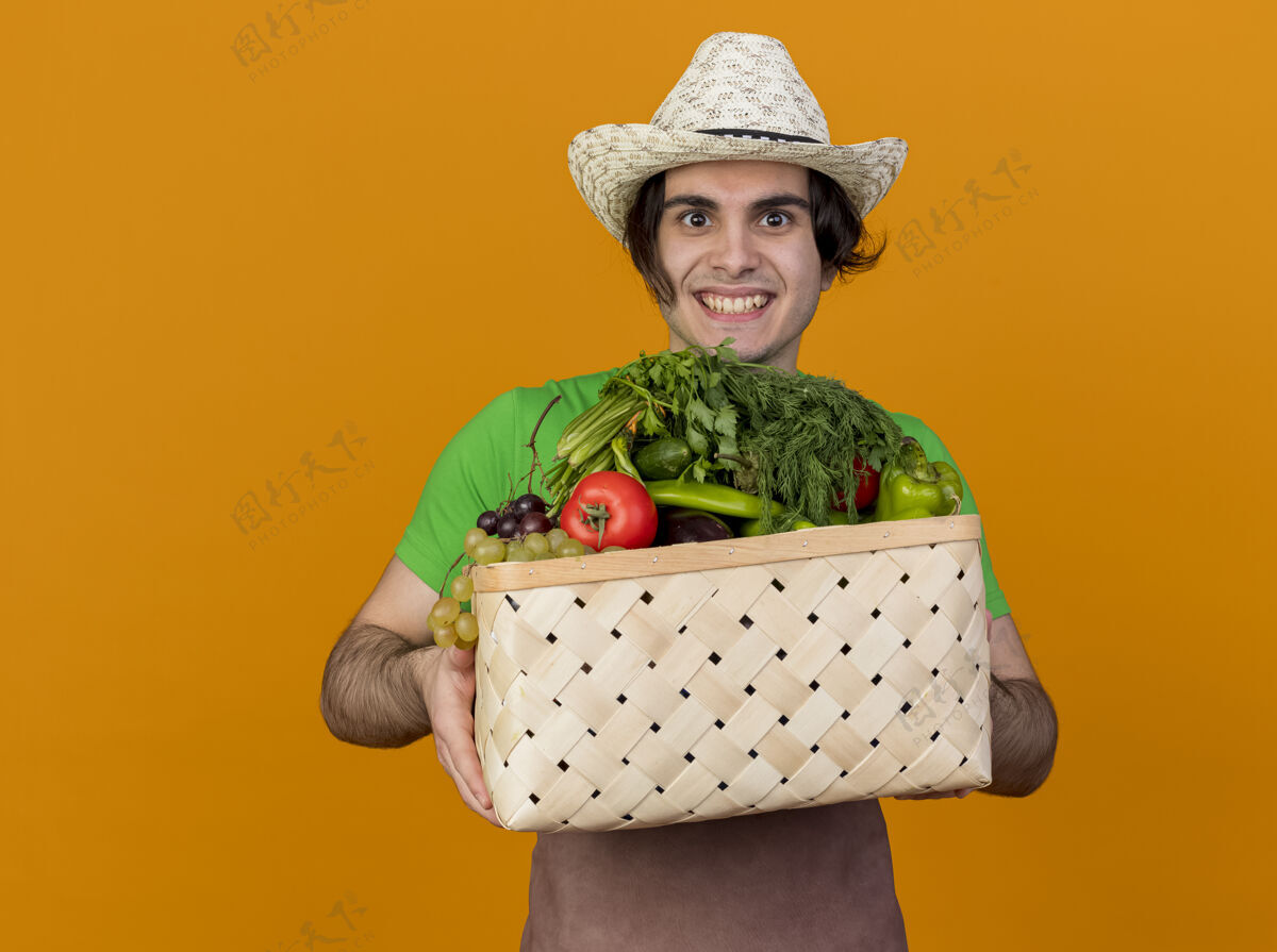 花园年轻的园丁 围着围裙 戴着帽子 手里拿着装满蔬菜的箱子 站在橙色的背景上 面带微笑地看着摄像机蔬菜立场举行