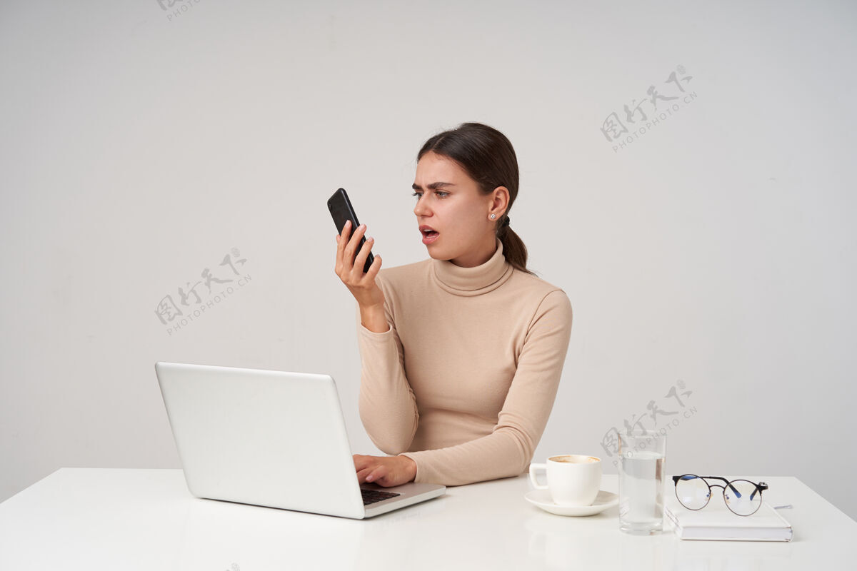 马尾辫室内拍摄的不高兴的年轻黑发女士看着手机 撅着嘴 做鬼脸 手放在键盘上 坐在白墙上坐着女士鬼脸