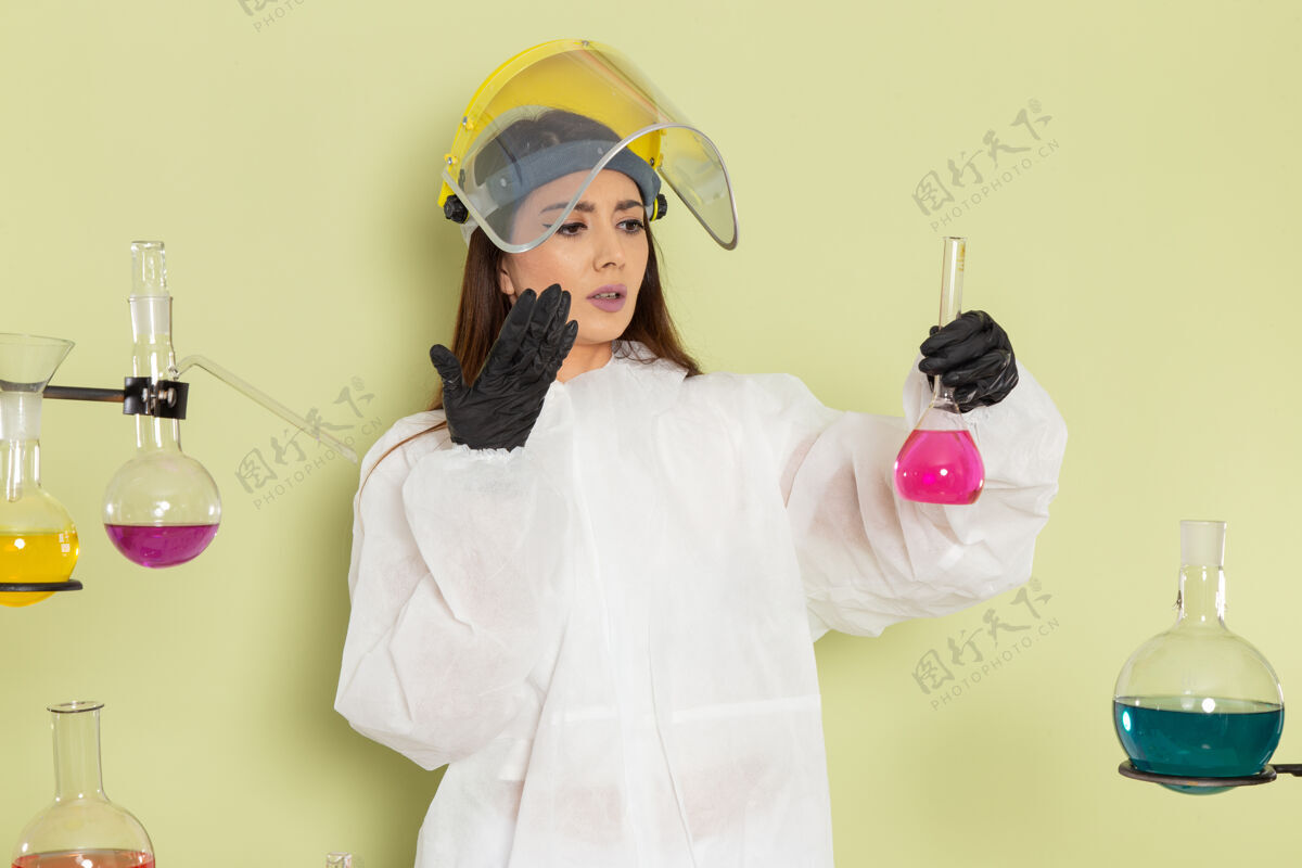 女正面图穿着特殊防护服的女化学家拿着绿色表面上有粉红色溶液的烧瓶工作化学家化学品
