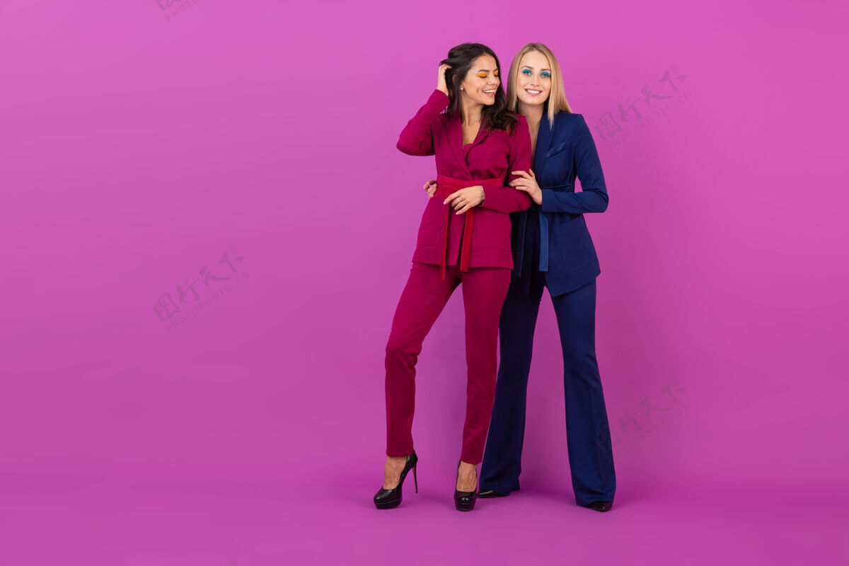 奢华高级时尚风格两位微笑迷人的女士在紫罗兰色的墙壁上穿着时尚多彩的紫色和蓝色晚礼服 朋友们一起玩 时尚潮流衣服人庆典