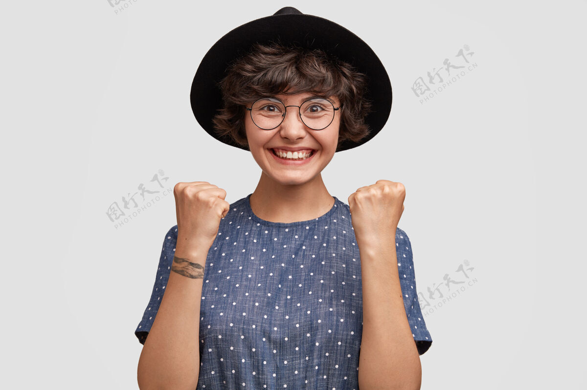 高兴快乐的白种人可爱的年轻女模特握紧拳头帽子水平眼镜