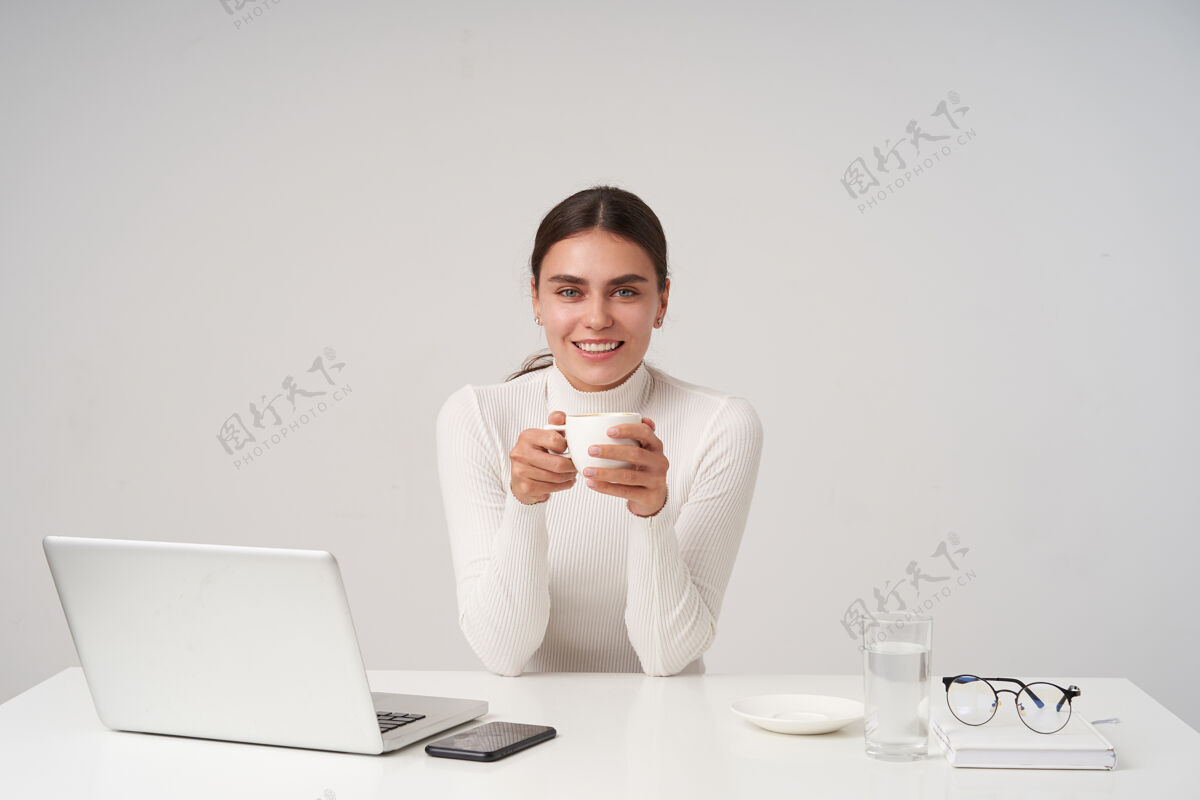 眼镜年轻开朗的蓝眼睛黑发女性 自然妆容 双手捧着一杯茶 对着镜头开心地微笑 在白墙上摆姿势头发桌子微笑