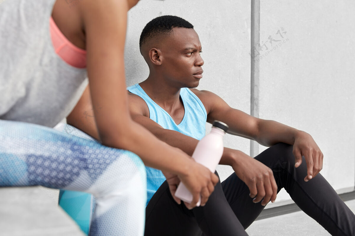 男孩皮肤黝黑的男性坐累了 他的伴侣拿着一瓶冷水在旁边摆姿势 一起做运动 过着积极的生活方式 经常在健身房或户外训练 穿着运动服非洲不规则剪裁男朋友