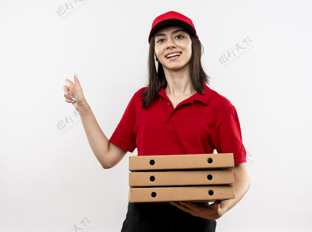 手指穿着红色制服 戴着帽子 拿着一叠披萨盒 用食指指着旁边 高兴地站在白色背景上微笑的年轻送货女孩送货帽子女孩