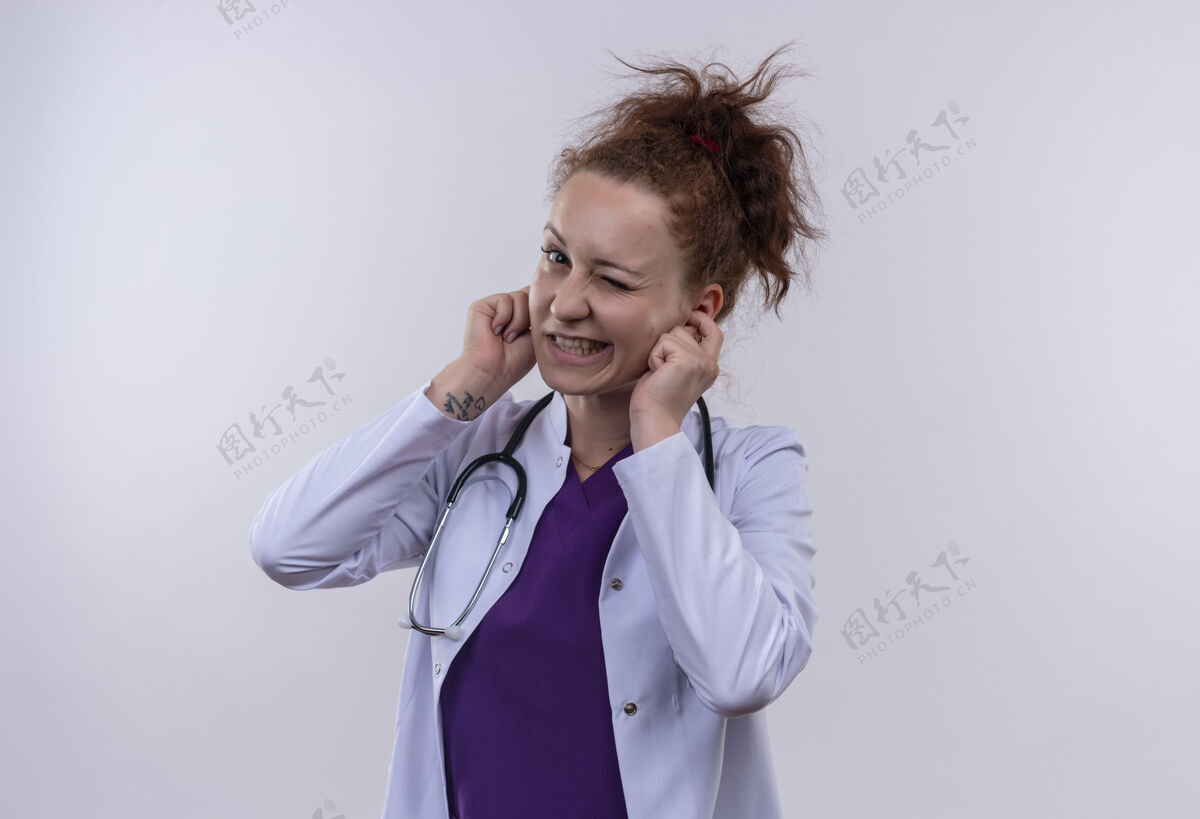 女人年轻的女医生穿着白大褂 带着听诊器 用手指闭着耳朵 脸上带着恼怒的表情 为站在白墙上的噪音表情外套噪音