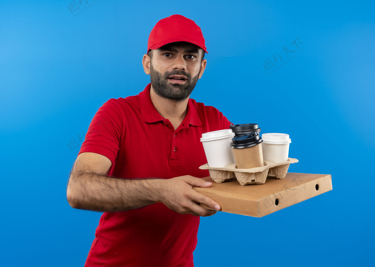 披萨站在蓝色的墙上 一个留着胡须 穿着红色制服 戴着帽子 手里拿着披萨盒和咖啡杯的送货员 看上去很困惑 很焦虑拿着非常看