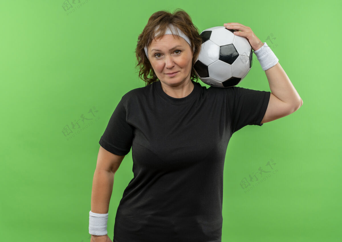老年身穿黑色t恤 头箍 手持足球 表情严肃自信的中年运动女性站在绿色墙壁上头带球表情
