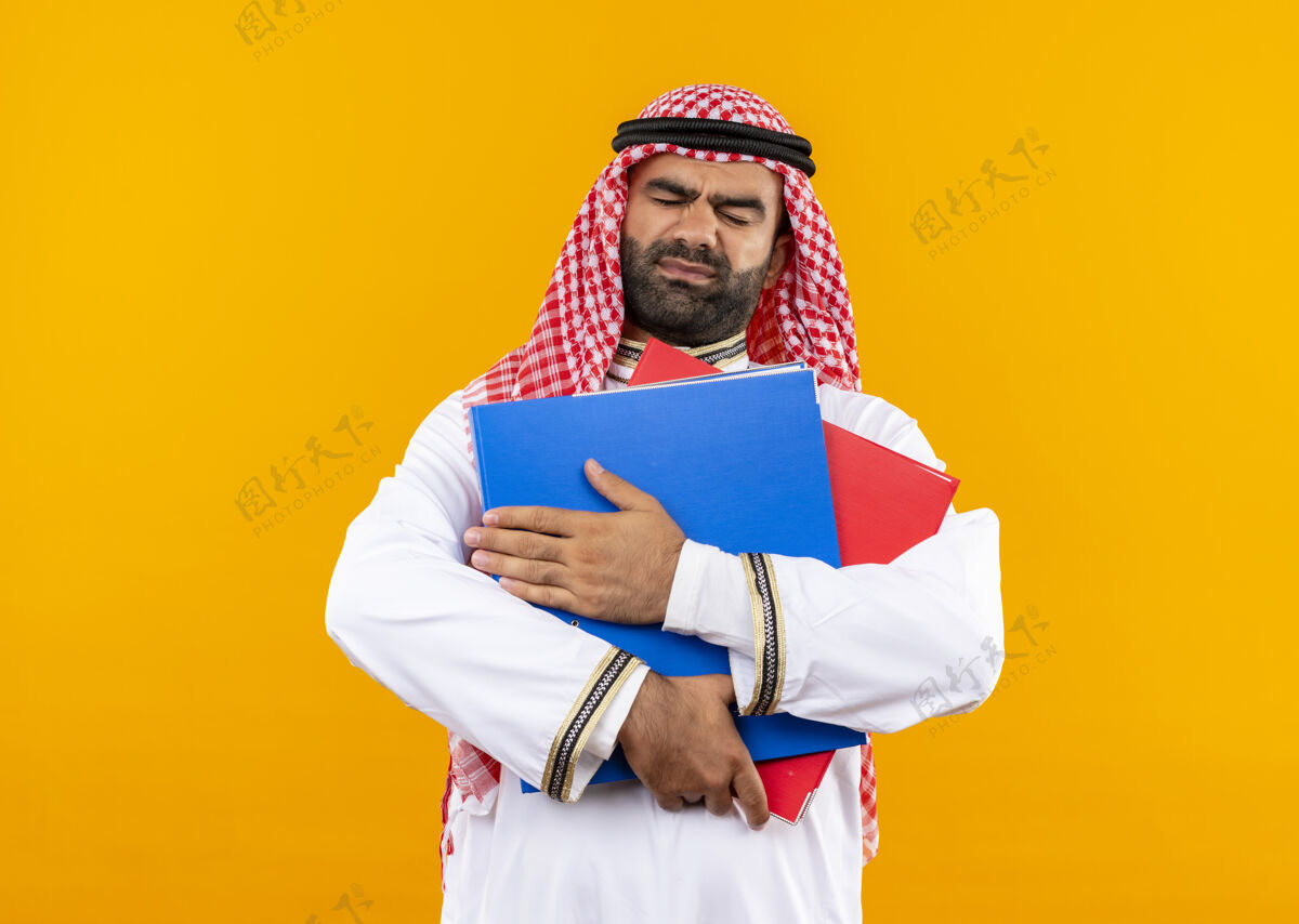 持有身着传统服装的阿拉伯商人站在橙色的墙上 手里拿着两个文件夹 看上去既不高兴又不高兴文件夹传统穿