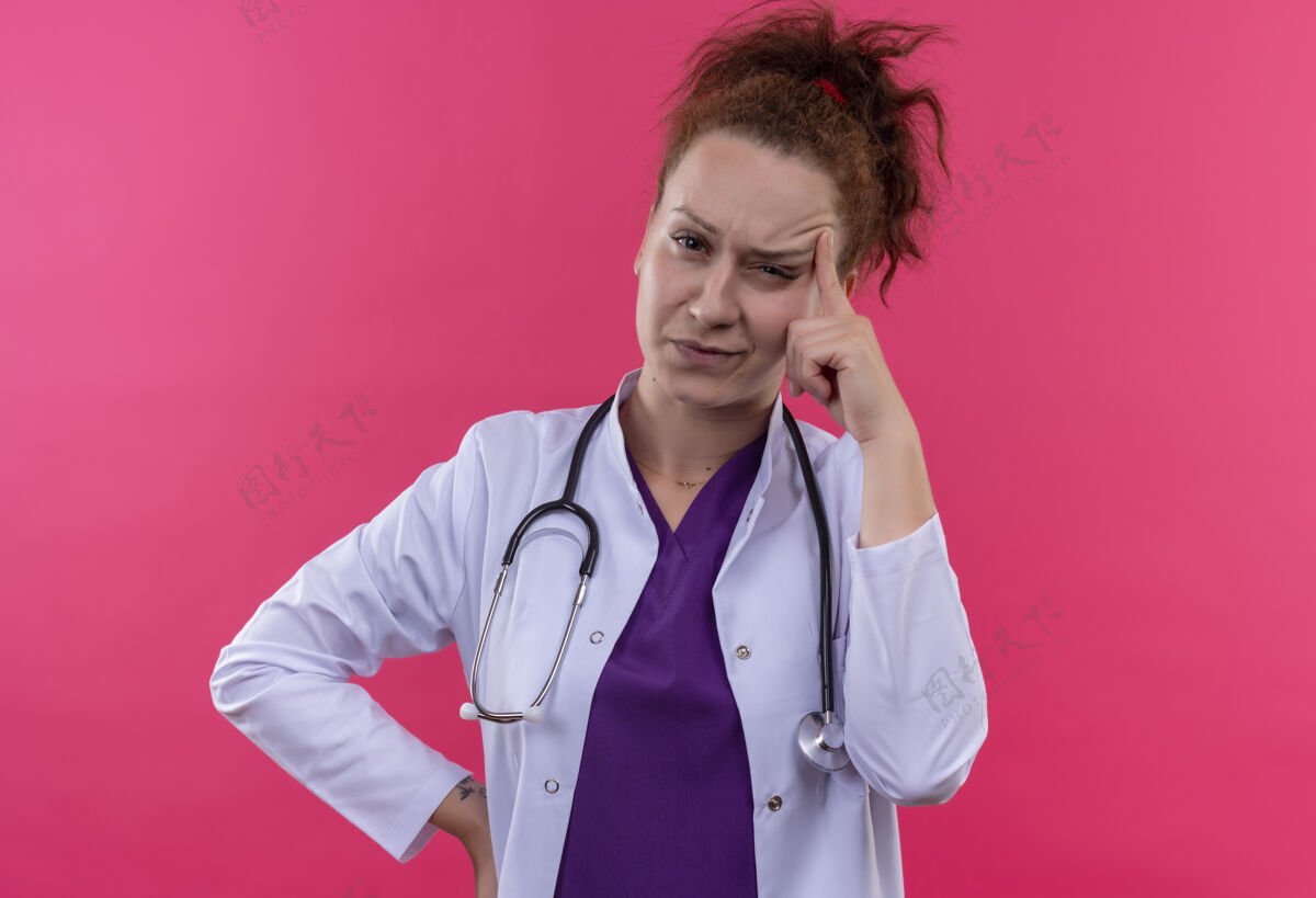 年轻年轻的女医生穿着白大褂 手持听诊器 带着怀疑的表情 指着站在粉红色墙上的太阳穴医生太阳穴看