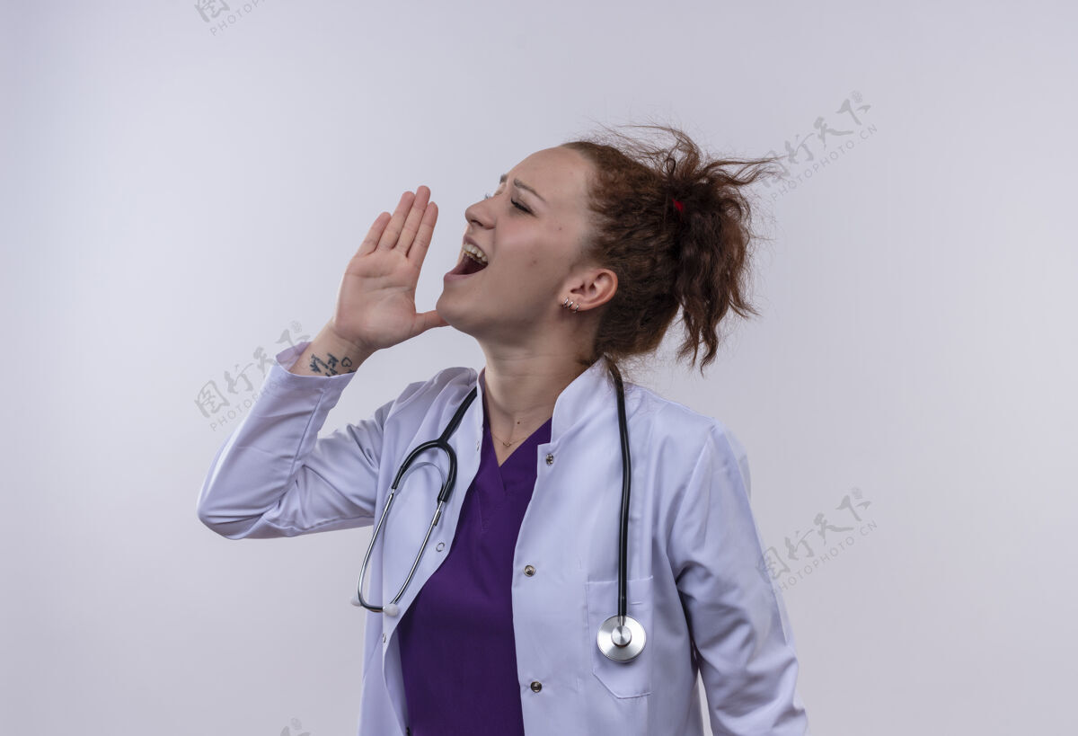 女人年轻的女医生穿着白大褂 手持听诊器 站在白墙上 用手捂着嘴大喊大叫手年轻人站着