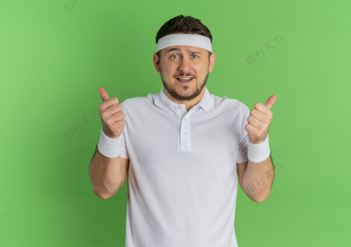 衬衫身穿白衬衫 头箍朝前微笑着竖起大拇指的年轻健身男子站在绿色的墙上头带男性运动