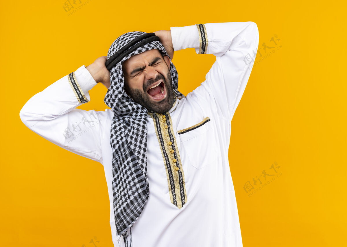 穿着阿拉伯商人穿着传统的疯狂服装 站在橙色的墙上 用咄咄逼人的表情抚摸着自己的手大声喊叫阿拉伯语疯狂传统