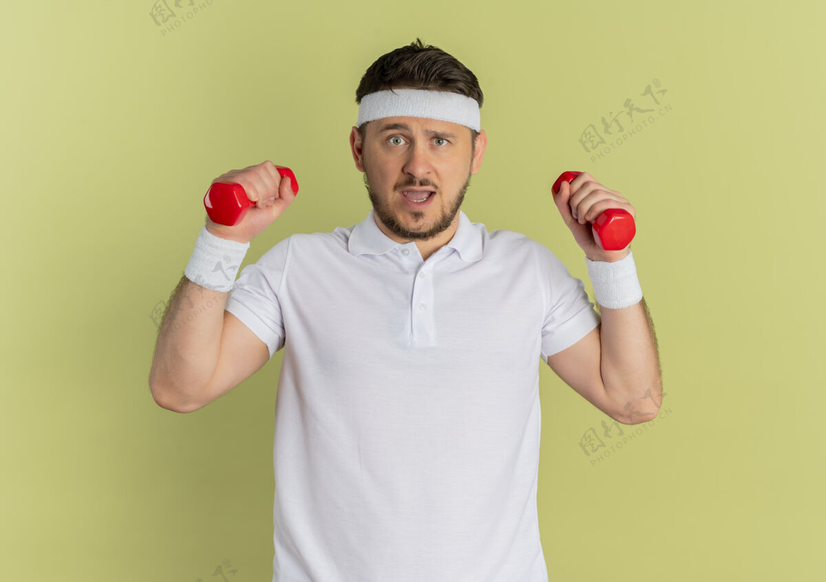 橄榄身穿白衬衫 头箍戴着哑铃的年轻健身男子站在橄榄墙上 看起来很困惑看运动运动装