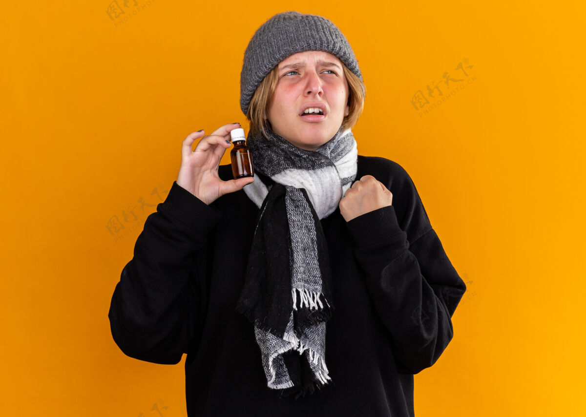 感觉不健康的年轻女子戴着暖和的帽子 脖子上围着围巾 感觉不舒服 患了感冒和流感 拿着药瓶 带着恼怒的表情看着橙色的墙上帽子穿生病