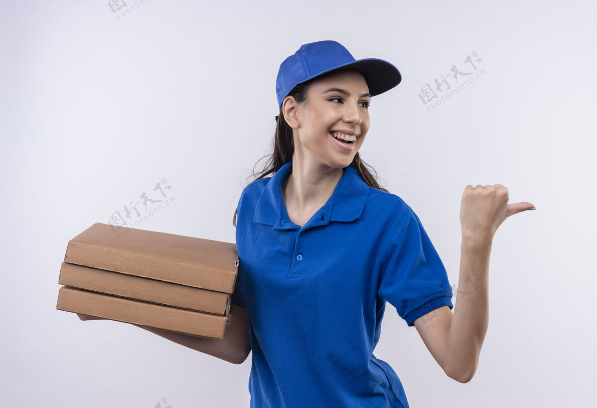 蓝色穿着蓝色制服 戴着帽子的年轻送货女孩拿着一叠披萨盒 拇指朝后 愉快地微笑着微笑女孩送货
