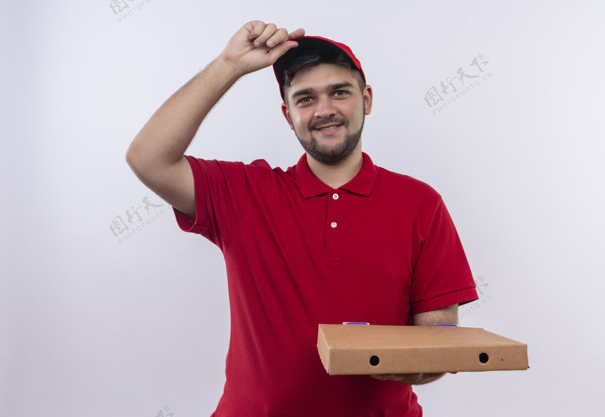 年轻年轻的送货员穿着红色制服 戴着帽子 手里拿着皮萨盒 看着相机 自信地微笑着抚摸着他的帽子盒子递送自信