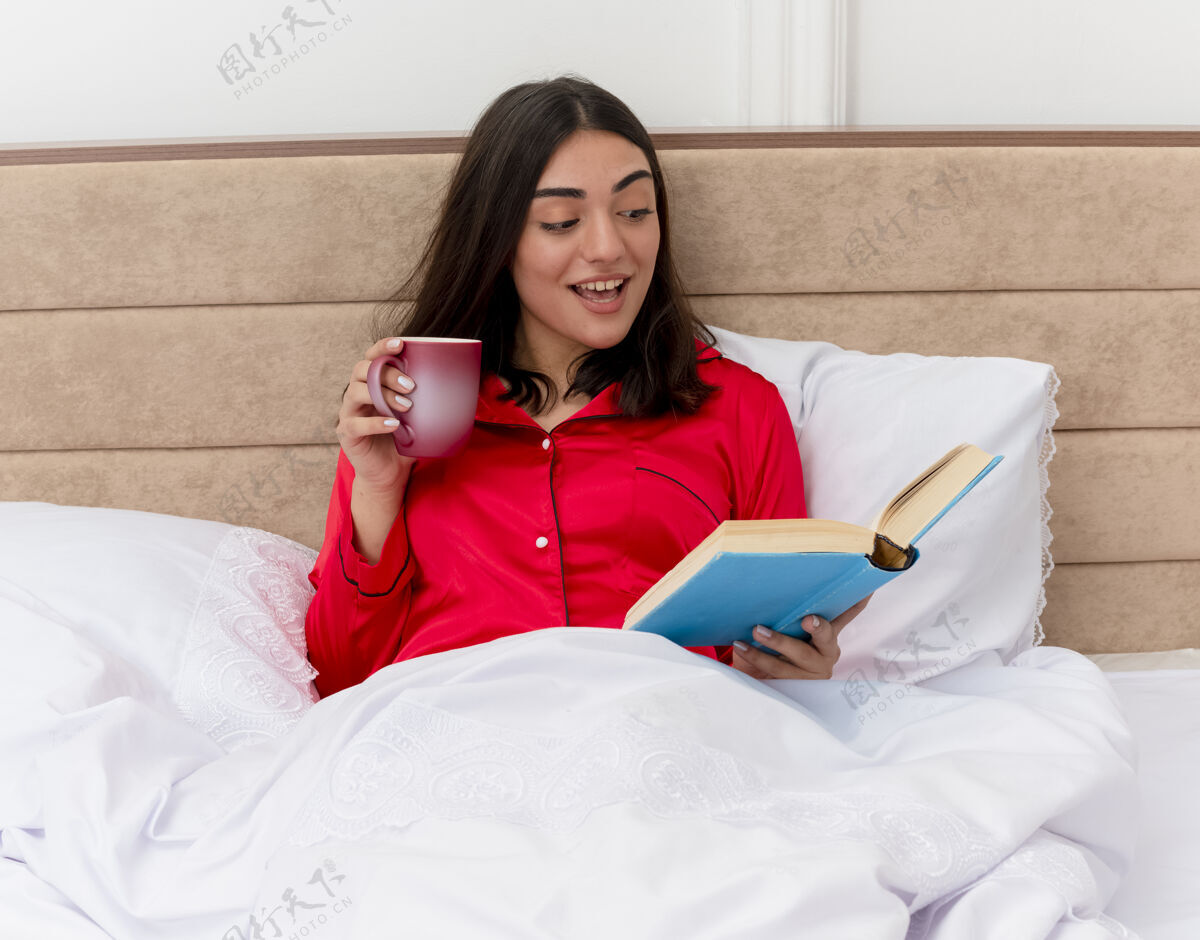 阅读穿着红色睡衣的年轻漂亮女人坐在床上 喝着咖啡 看书 放松地享受周末 微笑着 愉快地在卧室里读着书 背景很浅享受红色快乐