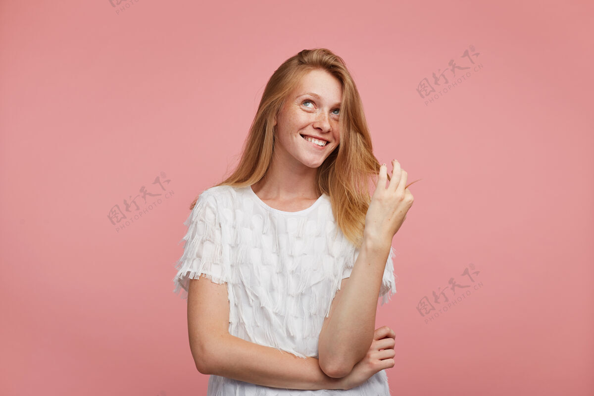 年轻快乐的年轻迷人的红发女性 抬起手拉着头发 带着幸福的笑容 梦幻般地向上看 站在粉色的背景上衬衫表情肖像