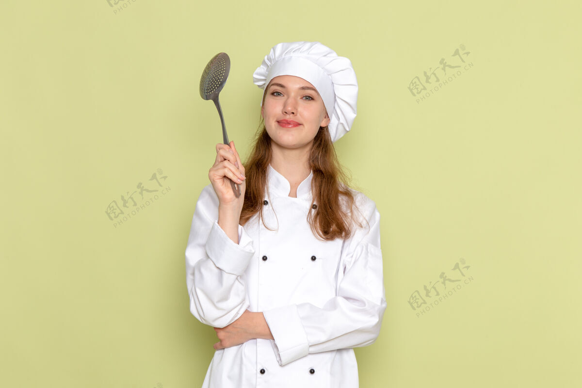 医生身穿白色厨师服的女厨师手持大银勺 微笑着站在绿色的墙上制服前面专业