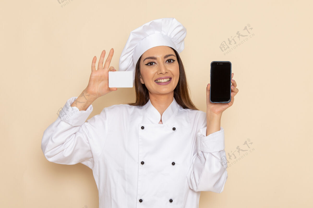 女性身穿白色厨师服的年轻女厨师的正视图 在浅白的墙上拿着名片和电话前面电话举行